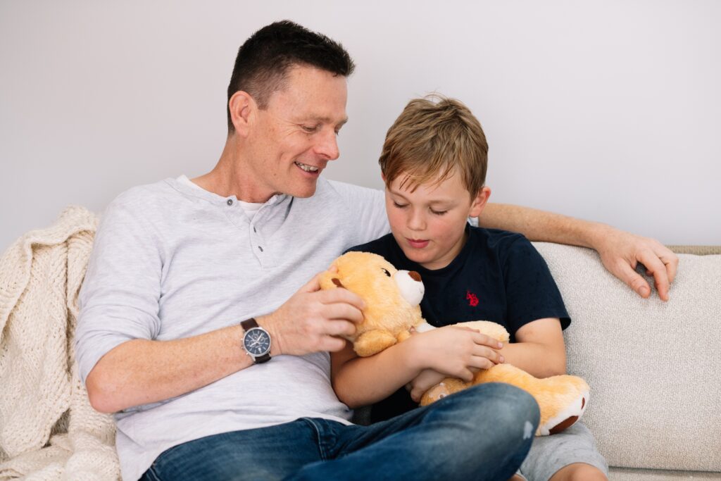 Father and son sitting on a couch talking together while holding a Tedology wellbeing bear