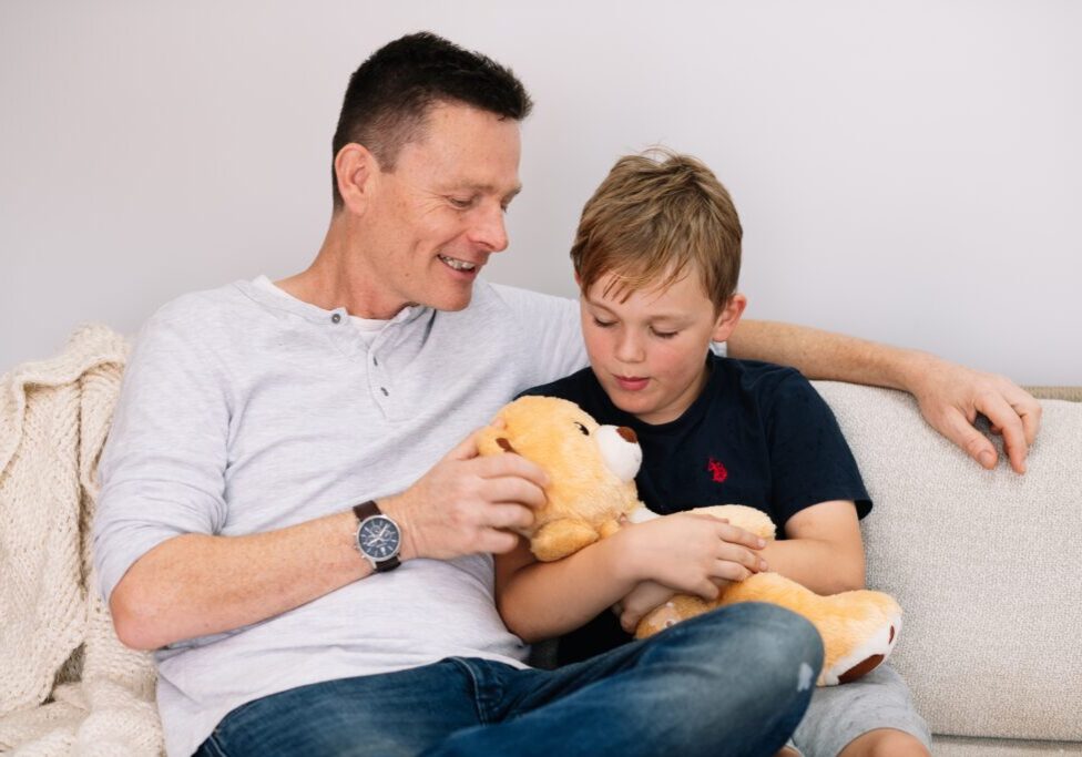 Father and son sitting on a couch talking together while holding a Tedology wellbeing bear